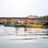 Zwei Personen paddeln entspannt in einem aufblasbaren Kajak in Marine-Gelb auf ruhigem Wasser, umgeben von einer malerischen Stadtlandschaft und einer alten Steinbrücke.