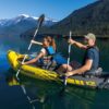 Zwei Personen paddeln in einem gelben aufblasbaren Kajak namens Explorer auf einem ruhigen See, umgeben von bewaldeten Bergen und blauem Himmel. Das Bild vermittelt Abenteuerlust und Naturverbundenheit.