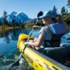Eine junge Frau paddelt in einem gelben aufblasbaren Kajak Explorer K2 auf ruhigem Wasser, umgeben von majestätischen Bergen und klarem blauen Himmel während eines Outdoor-Abenteuers.