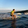 Männlicher Wassersportler mit leuchtend grüner Schwimmweste auf einem Surfbrett, der hinter einem Motorboot gezogen wird, umgeben von einer malerischen Wasserlandschaft bei Sonnenuntergang.