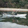 Mann paddelt in einem weißen faltbaren Kajak auf ruhigem Wasser, umgeben von Ufervegetation und sanften Hügeln, ideal für Freizeitpaddeln.